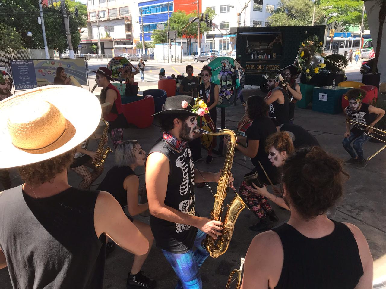 Apresentação fanfarra Cumbia Calavera embaixo do viaduto, integrantes vestidos de preto e maquiagem caracterizada de caveira mexicana